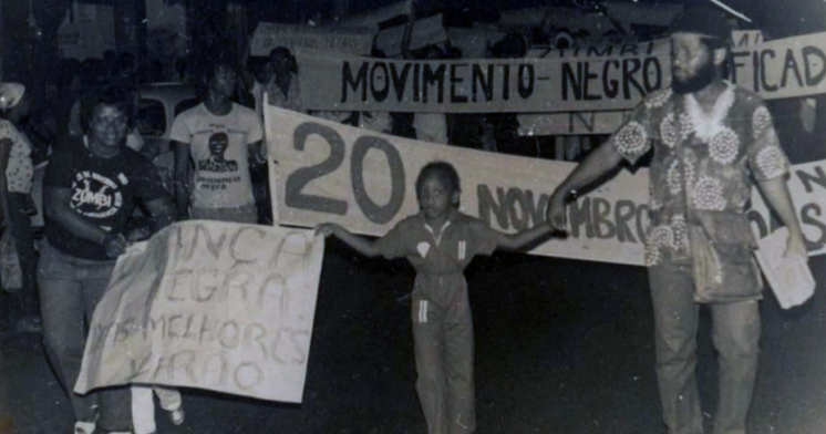 pessoas negras, segurando cartazes e faixas com inscrição "Movimento Negro Unificado" e "20 de novembro"