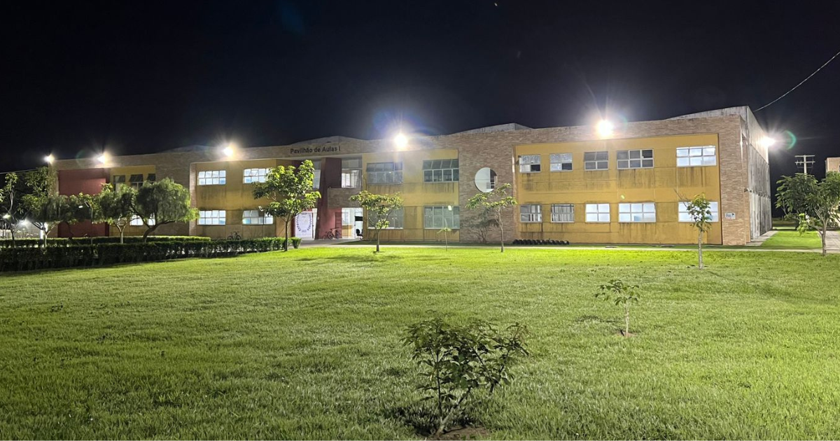 A imagem mostra um prédio de dois andares à noite, iluminado por várias luzes externas. O edifício tem uma fachada de tijolos e partes pintadas de amarelo e vermelho. Na parte superior central do prédio, há uma placa com os dizeres "Pavilhão de Aulas I". Em frente ao prédio, há um gramado bem cuidado com algumas árvores pequenas e arbustos. A iluminação é forte, destacando tanto o prédio quanto o gramado à sua frente.
