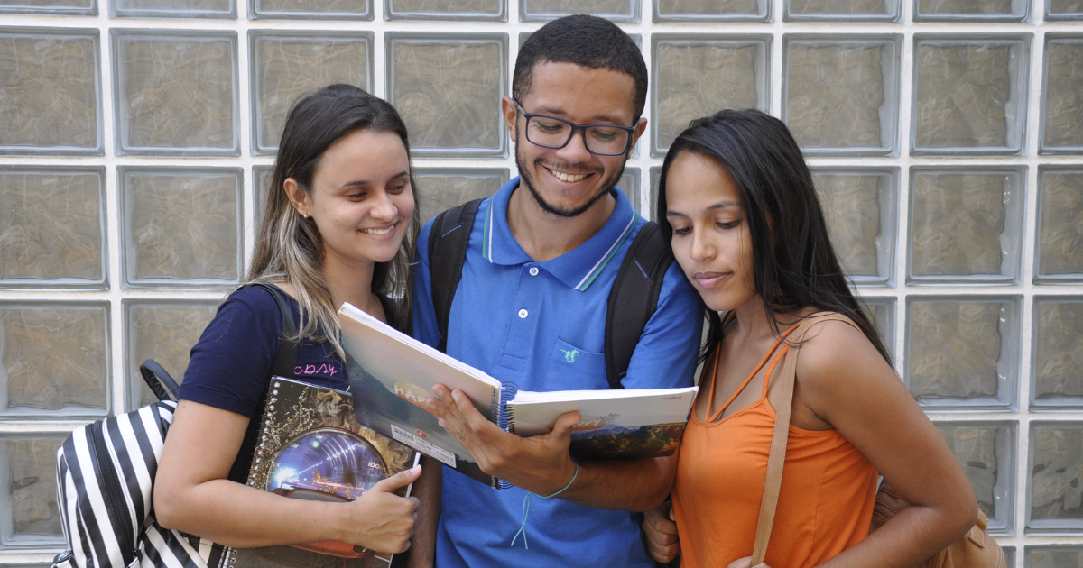 A imagem mostra três jovens estudantes sorrindo e olhando para um livro que estão segurando juntos. Eles estão em frente a uma parede de blocos de vidro. A jovem à esquerda tem cabelo castanho claro, veste uma camiseta preta e carrega uma mochila listrada. O jovem no meio usa óculos, tem cabelo curto e barba rala, veste uma camisa polo azul e carrega uma mochila preta. A jovem à direita tem cabelo longo e escuro, veste uma blusa laranja e também carrega uma mochila, Todos parecem estar felizes e concentrados no livro.