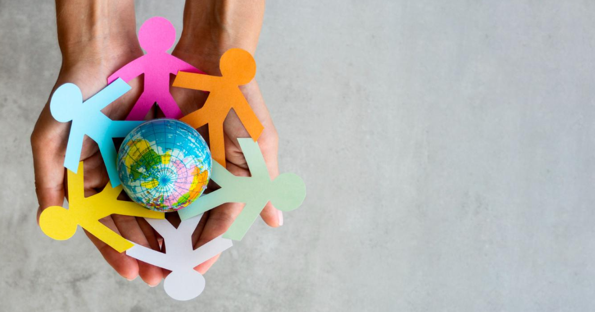 A imagem mostra duas mãos segurando um círculo de figuras humanas de papel colorido, que estão de mãos dadas. No centro do círculo, há um pequeno globo terrestre. As figuras de papel são de várias cores, incluindo rosa, laranja, verde, azul e amarelo, representando diversidade e união. O fundo da imagem é cinza e liso.