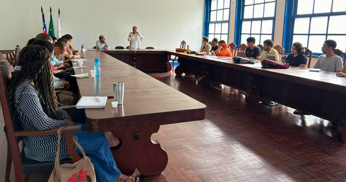 A imagem mostra uma sala de reuniões com uma mesa grande em formato de U. Há várias pessoas sentadas ao redor da mesa, participando de uma reunião. A maioria das pessoas está olhando para uma mulher em pé no centro da sala, que parece estar falando ou apresentando algo. A sala tem janelas grandes à direita, permitindo a entrada de bastante luz natural. No fundo, há três bandeiras: a do Brasil, a da Bahia e a da UFRB. As pessoas estão vestidas de maneira casual e há diversos itens sobre a mesa, como garrafas de água, cadernos e canetas.