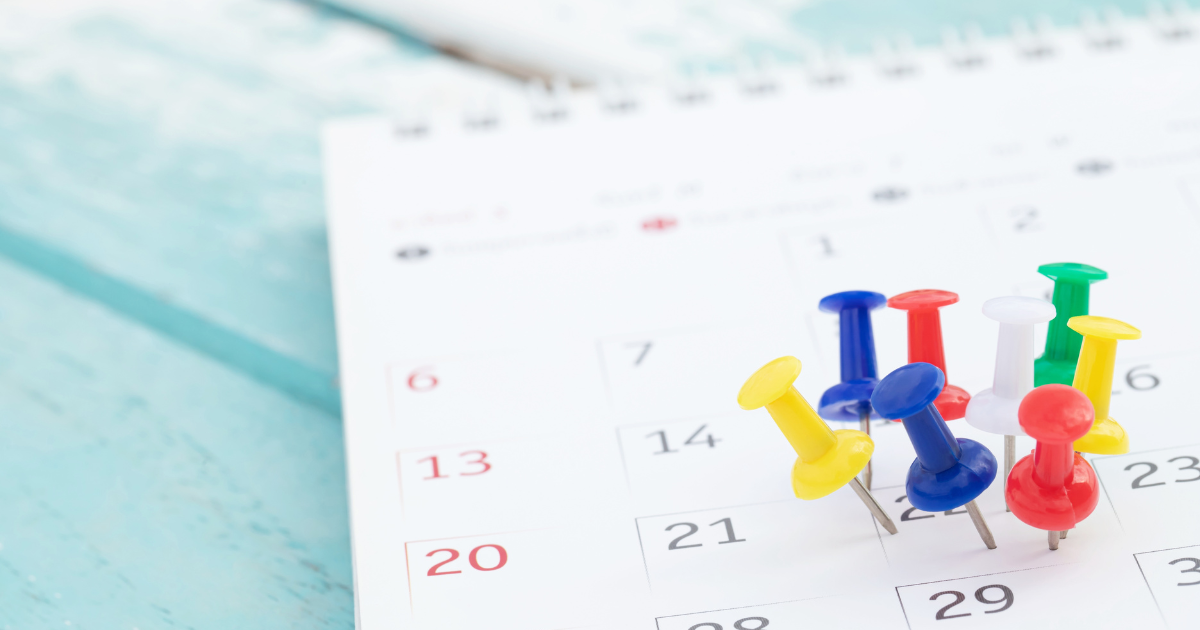 A imagem mostra um calendário de papel com vários alfinetes coloridos espetados nele. Os alfinetes são de cores variadas, incluindo amarelo, azul, vermelho, verde e branco. O calendário está sobre uma superfície de madeira pintada de azul claro. As datas visíveis no calendário incluem os dias 6, 13 e 20, que estão destacados em vermelho, indicando que são domingos.