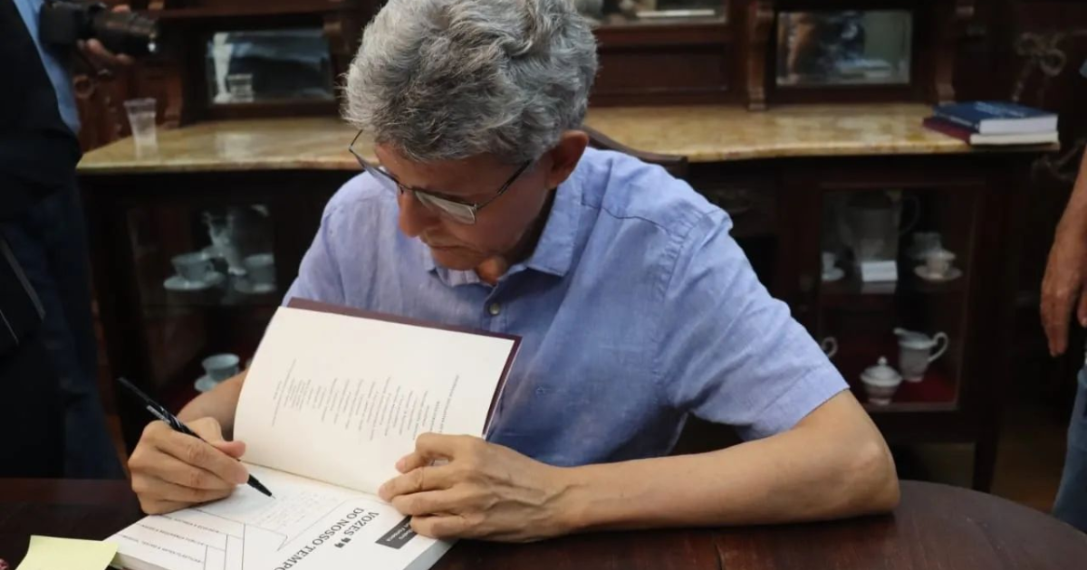 A imagem mostra um homem de cabelos grisalhos e curtos, usando óculos e uma camisa azul clara, sentado em uma mesa de madeira. Ele está autografando um livro com uma caneta preta. O ambiente parece ser uma livraria ou biblioteca, com prateleiras de madeira ao fundo, onde há xícaras e pires expostos. Há também um copo plástico transparente sobre a mesa ao fundo.