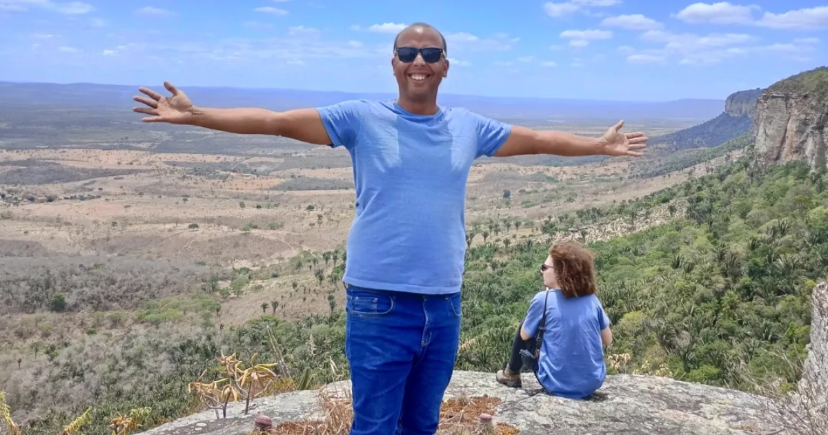 A foto mostra duas pessoas em um cenário natural. Em primeiro plano, há um homem de pele escura, usando óculos de sol, camiseta azul clara e calça jeans azul. Ele está de pé, com os braços abertos e um sorriso no rosto. Ao fundo, sentada em uma rocha, há uma mulher de pele clara, cabelos castanhos e cacheados, também vestindo uma camiseta azul clara e calça escura. Ela está de costas para a câmera, olhando para a paisagem. A paisagem ao redor é composta por um vasto vale com vegetação esparsa e algumas áreas verdes, além de montanhas ao fundo sob um céu azul com algumas nuvens.