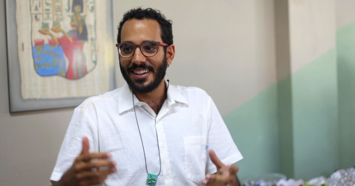 A imagem mostra um homem sorridente com barba e cabelo curto e encaracolado. Ele está usando óculos de armação escura e uma camisa branca de manga curta. 0 homem também está usando um colar com um pingente verde. Ele parece estar gesticulando com as mãos enquanto fala, Ao fundo, há uma parede com um quadro que parece ter uma arte colorida, possivelmente com elementos culturais ou históricos. A parede tem uma cor clara, com uma faixa verde na parte inferior.