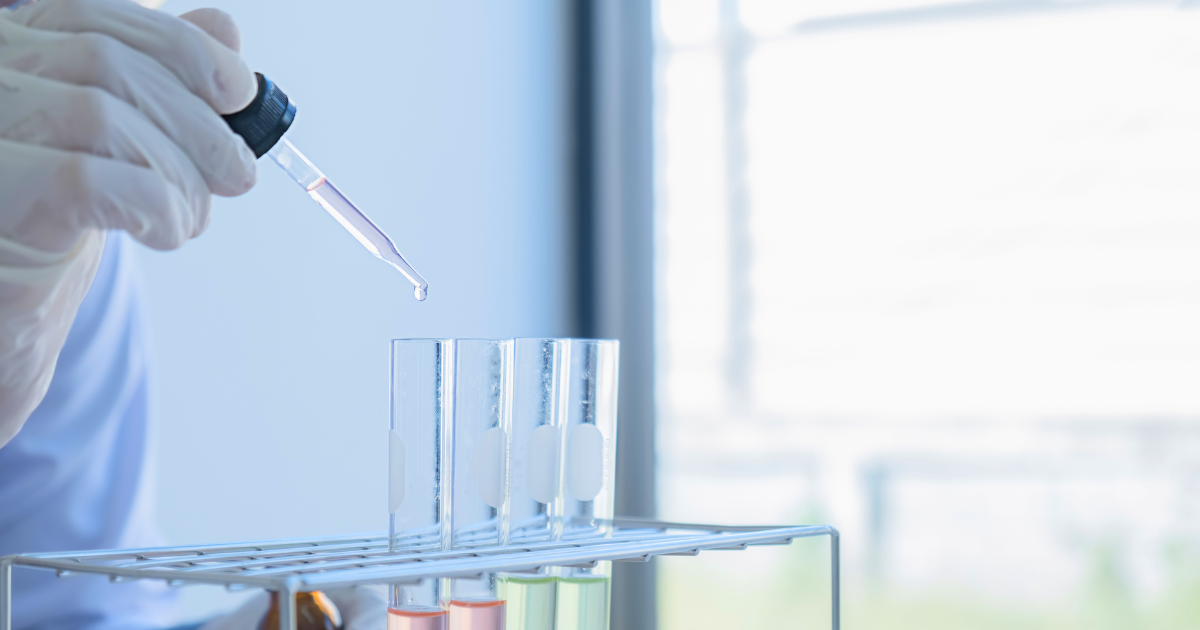 A imagem mostra uma mão usando luvas de látex segurando um conta-gotas, de onde está caindo uma gota de líquido em um tubo de ensaio. Há três tubos de ensaio em um suporte de metal, e cada um contém um líquido de cor diferente. O fundo é desfocado, sugerindo que a cena está em um laboratório.